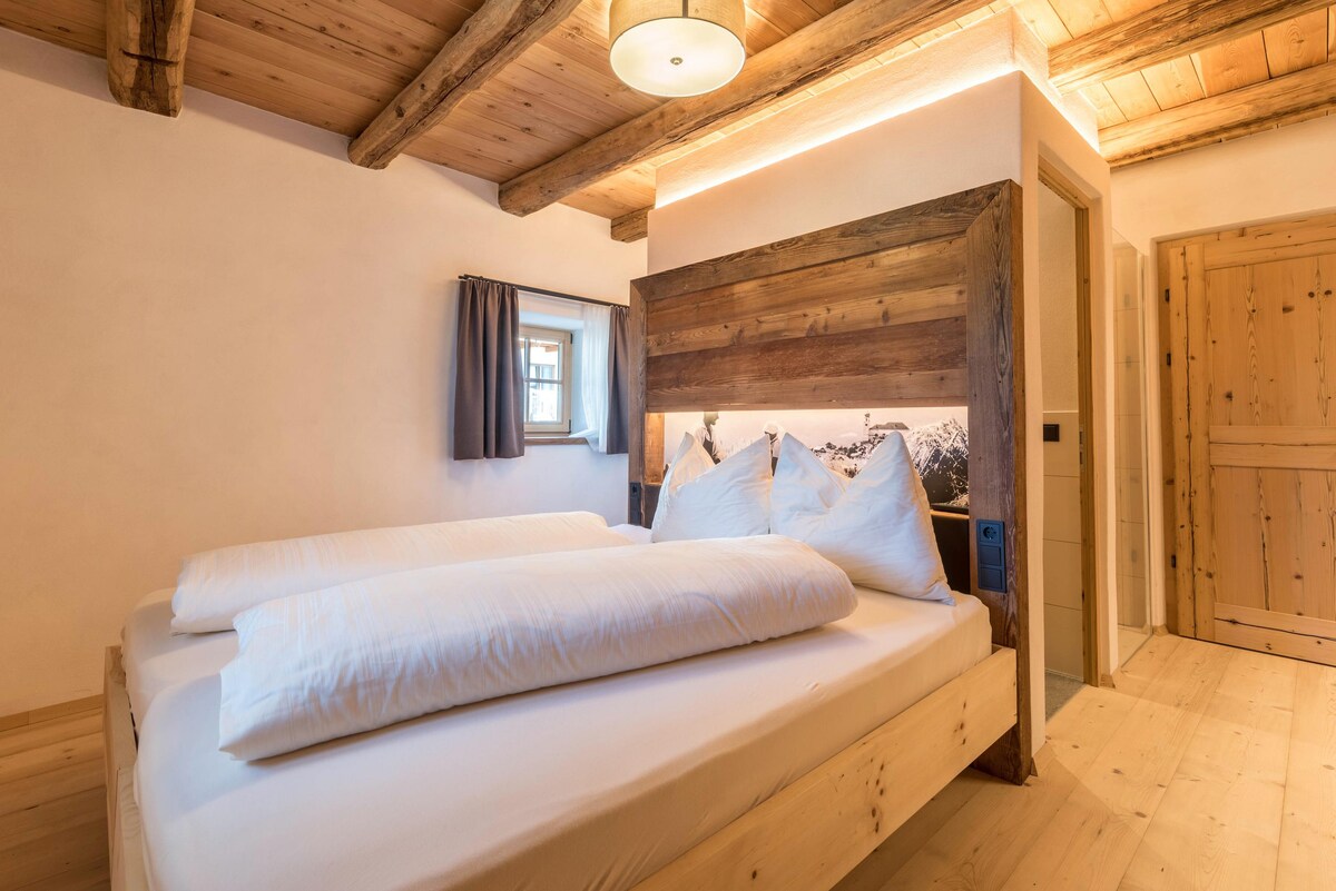 A cozy sleeping area is presented with two spacious beds, accompanied by large, soft white comforters and pillows. Wooden beams and a warm wood panel highlight the ceiling and wall, while a window with a curtain allows natural light to enter the room.