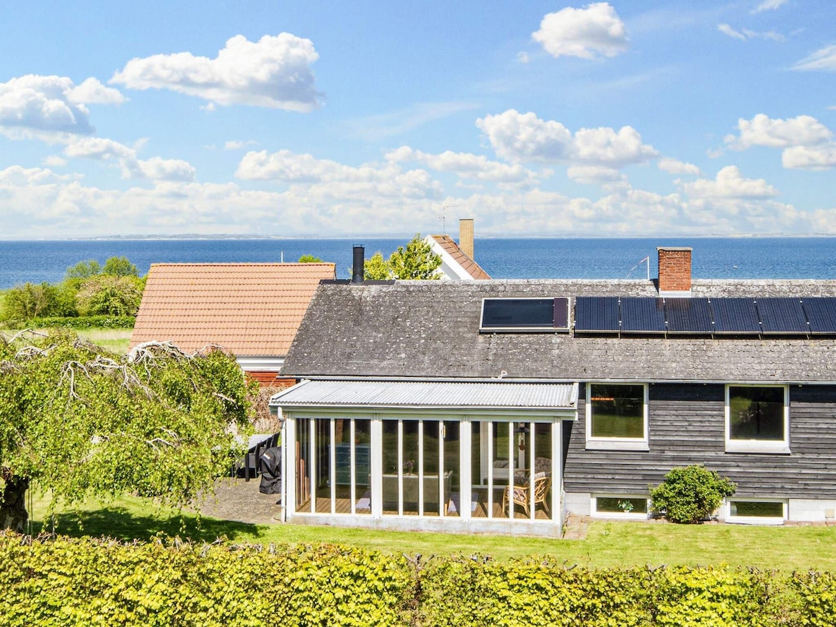 A view of the holiday home from a distance shows a sloped roof with solar panels. The house features large glass doors leading to the conservatory, set against a backdrop of lush greenery and the distant sea. A nearby house and a clear blue sky are also visible.