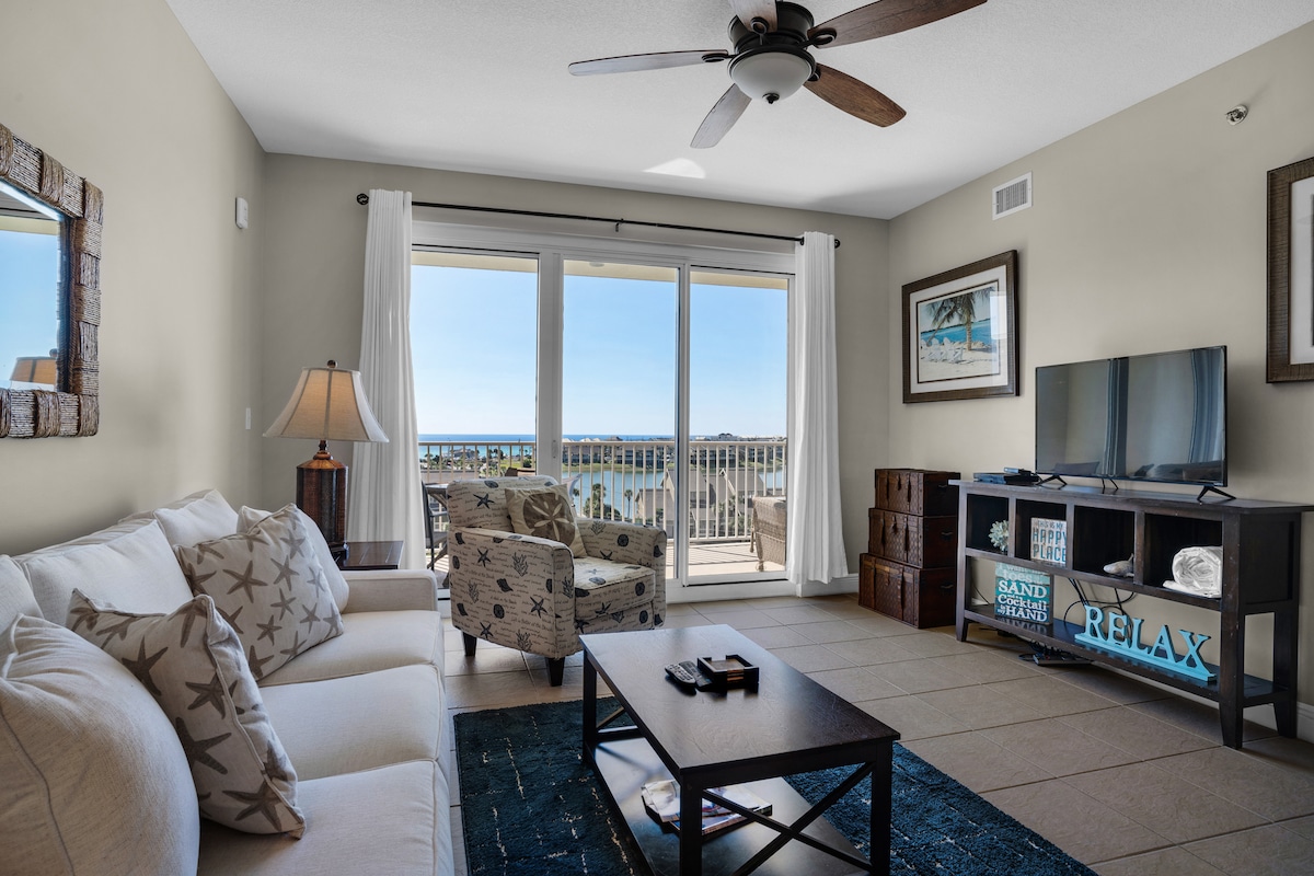 The living area features a comfortable couch paired with a decorative accent chair, a wooden coffee table, and a TV console with a flat screen TV. Large sliding glass doors open to a balcony offering views of the gulf and surrounding landscapes.