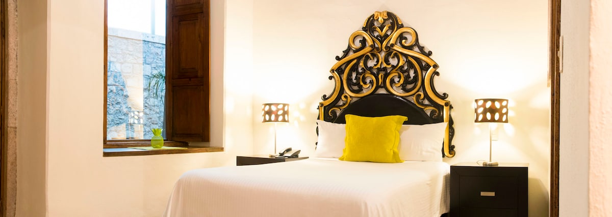 A well-appointed bedroom is depicted, featuring a spacious bed with an ornate headboard. Soft lighting is provided by bedside lamps, and a vibrant yellow pillow adds a pop of color. A wooden window frame opens to an exterior view, enhancing the warm ambiance.