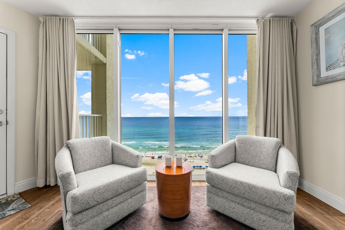 A view from the living area features two armchairs positioned around a round wooden table. Large floor-to-ceiling windows offer expansive views of the beach and ocean, with blue skies and fluffy white clouds in the background.