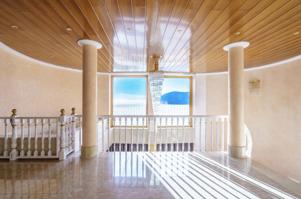An open and airy hallway features elegant columns and large windows that allow natural light to fill the space. A polished floor reflects the soft glow from the overhead chandelier, creating a bright and inviting atmosphere. Sweeping views of the surrounding landscape are visible through the windows.