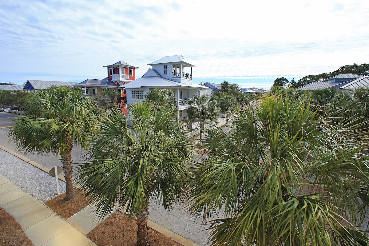 The image presents a view of a multi-story beach home surrounded by palm trees. The architecture features a mix of colors and shapes, with a hint of the ocean visible in the distance. The ground is covered with crushed stone, complementing the tropical environment.