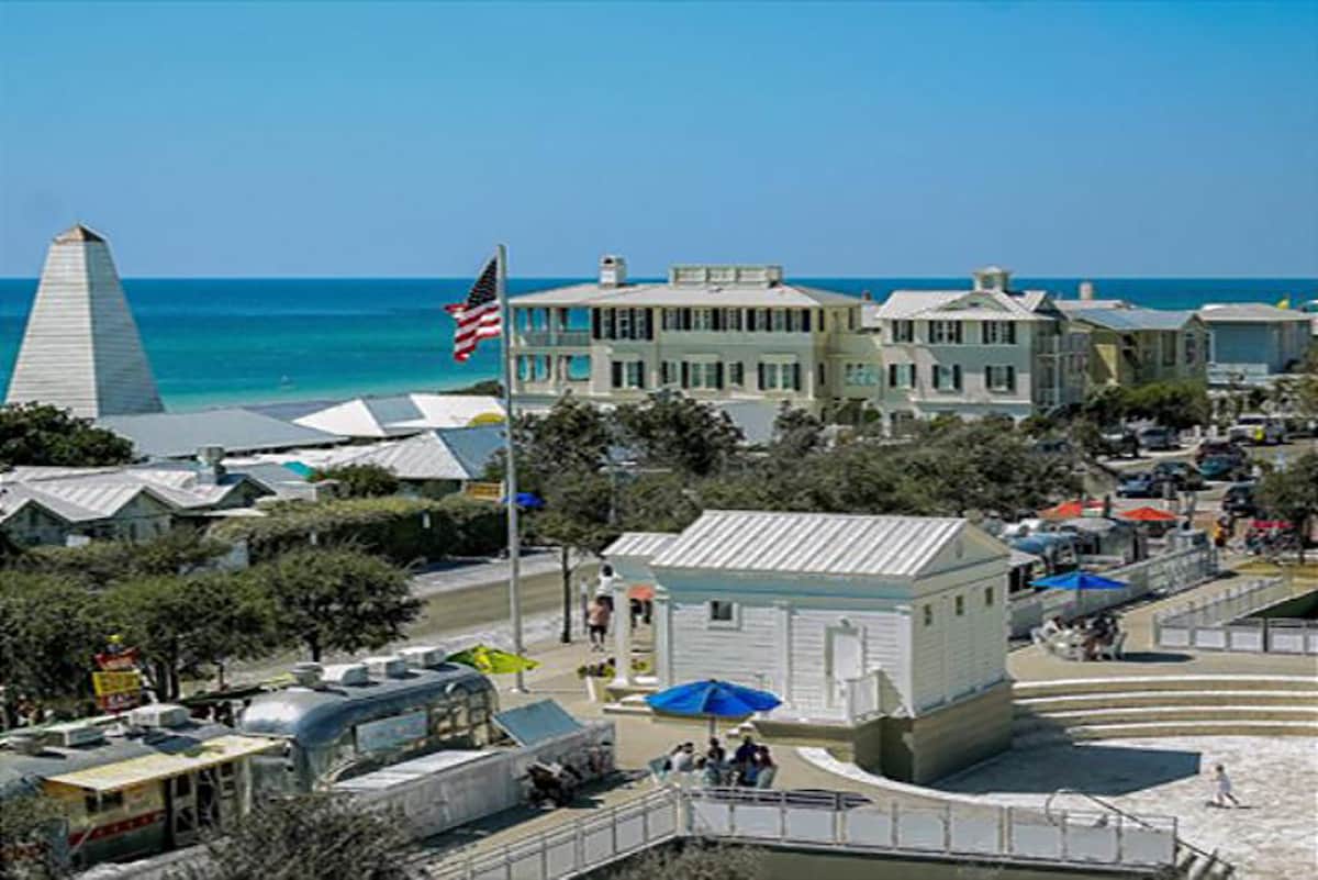 A coastal scene of Seaside, featuring a view of the beach and ocean under a clear blue sky. The image includes traditional wood-framed cottages, a prominent flag pole with an American flag, and people enjoying the vibrant atmosphere along the walkway.