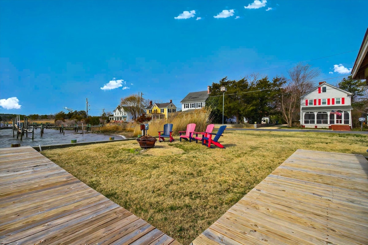 A spacious yard is shown with colorful adirondack chairs arranged in a welcoming manner. The grassy area is bordered by a wooden deck that leads to the water, with several houses visible in the background, including one with a screened porch and red accents.