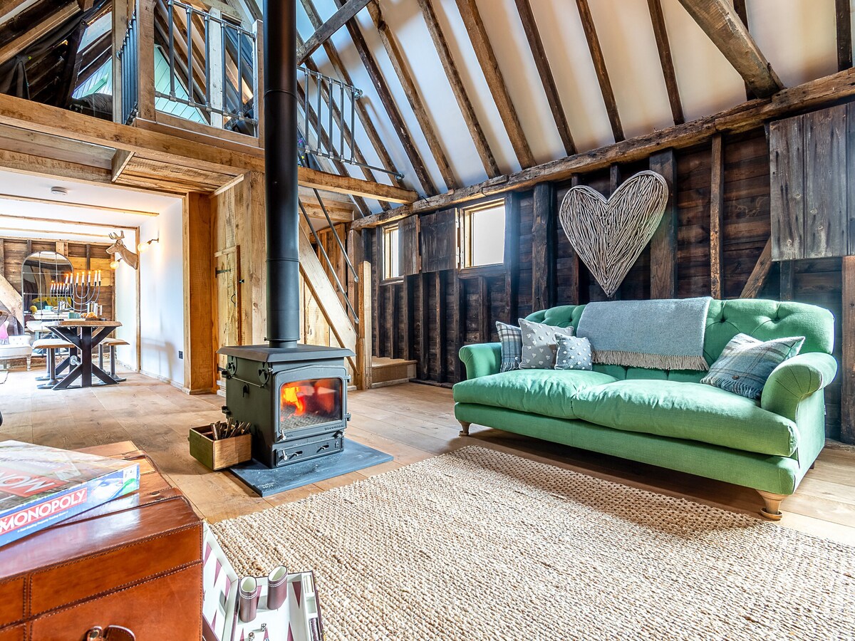 The living area features a vibrant green sofa positioned near a wood burner, providing warmth and comfort. Exposed wooden beams accent the ceiling, while light filters through multiple windows. A rustic wooden dining table is visible in the background, enhancing the inviting space.