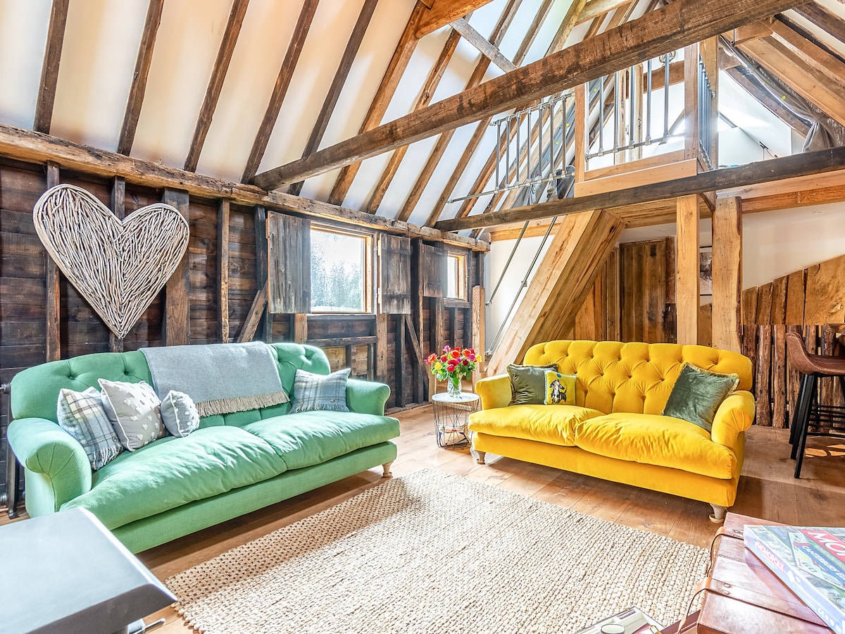 The living area features two vibrant sofas, one in green and another in yellow, set against rustic wooden walls with exposed beams. A natural fiber rug covers the wooden floor, complemented by a metal coffee table. Large windows allow ample natural light, enhancing the welcoming ambiance.