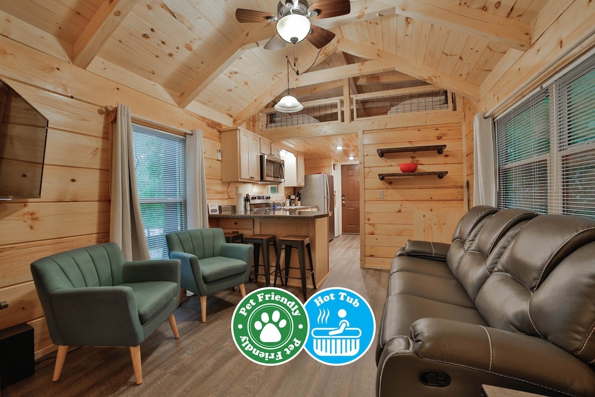 An inviting living area features a vaulted ceiling with wooden beams. Two green armchairs are positioned beside a black sofa, creating a cozy seating arrangement. The fully equipped kitchen is visible, showcasing wooden cabinetry and modern appliances. Natural light enters through window treatments.