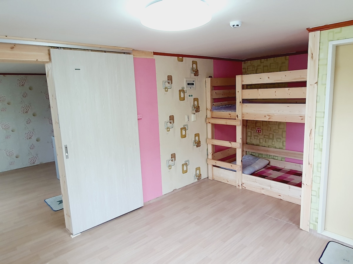 A bright room features light wooden floors and walls adorned with soft-patterned wallpaper. A wooden bunk bed is situated against a pink accent wall, with neatly arranged bedding inviting comfort. A sliding door leads to another area, enhancing the room's spaciousness.