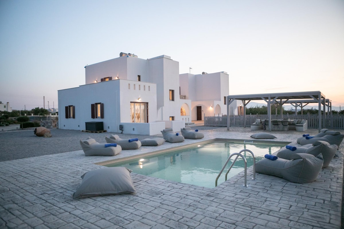 An inviting outdoor area features a swimming pool surrounded by comfortable lounge cushions. The villa, designed in a traditional Cycladic style, showcases whitewashed walls and large windows. A pergola offers shade, while the serene landscape provides a tranquil backdrop during twilight.