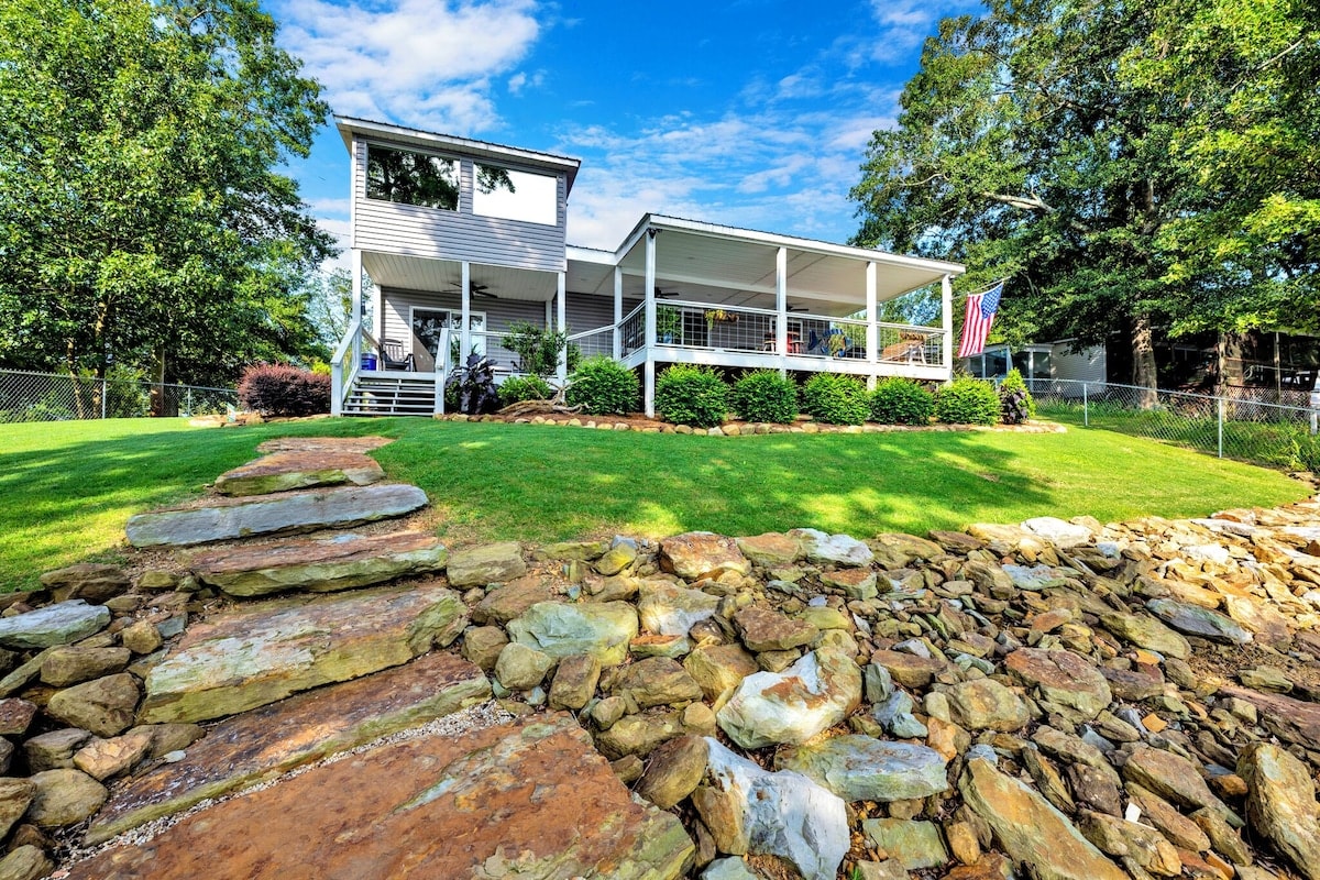 A two-story modern bungalow is situated beside a lush green yard, showcasing a well-maintained stone pathway leading to the water. A covered porch wraps around part of the house, with an American flag gently fluttering in the breeze against a backdrop of trees.