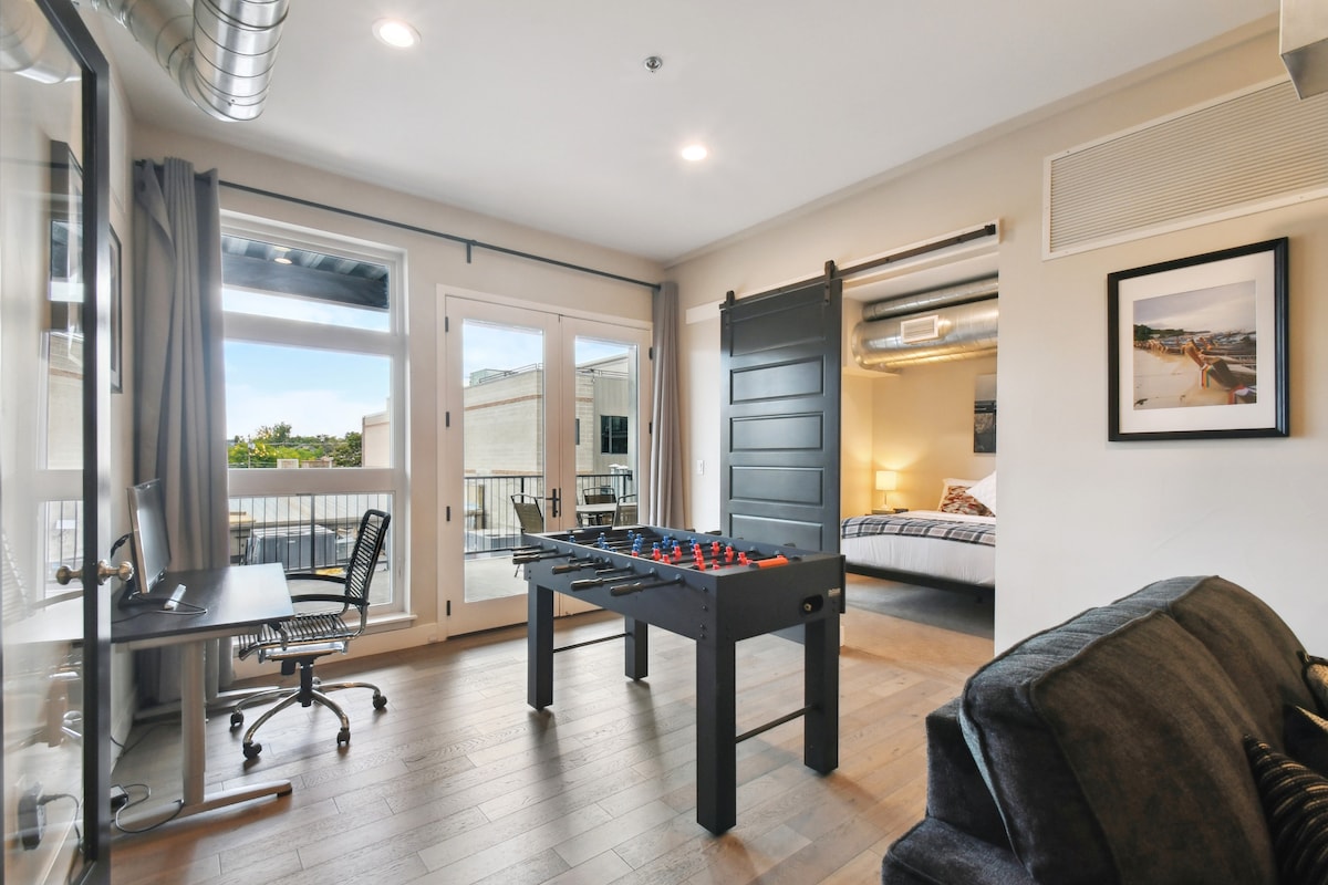 A versatile living space features a foosball table placed centrally. Natural light floods through floor-to-ceiling windows, revealing a view of the balcony. A black sliding barn door leads to the bedroom, while a desk and chair are positioned against the wall.