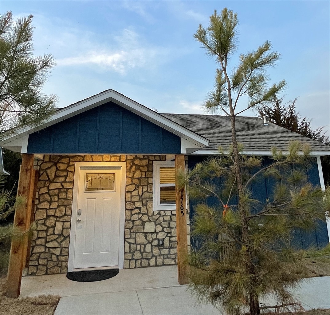 A cozy cottage exterior is showcased, featuring a stone and blue wooden facade. The front door is framed by a small window, with a welcoming mat positioned in front. A young pine tree is visible in the foreground, complementing the tranquil setting.