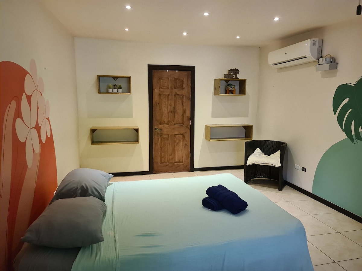 A spacious room features a large bed with soft linens and two dark towels neatly arranged on it. Shelves are mounted on the walls, holding decor and storage items. A wooden door leads outside, and an air conditioning unit is positioned above the bed.
