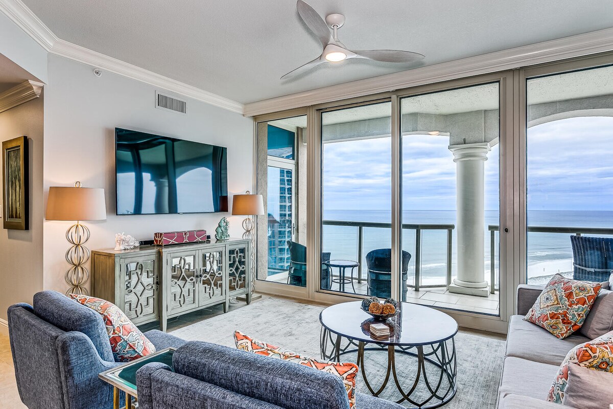 A spacious living area features large windows providing a panoramic view of the ocean. Comfortable seating arrangements include a stylish coffee table and patterned cushions. A sleek television is mounted on the wall, enhancing the sense of modern living, while natural light floods the room.