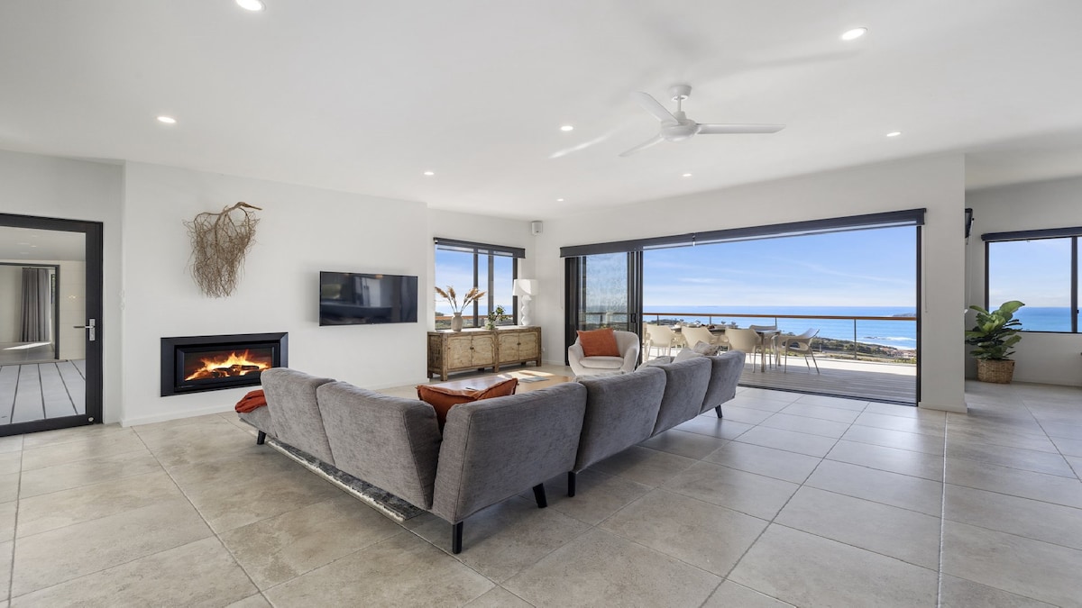 A spacious living area features large windows offering expansive ocean views. A comfortable grey sectional couch is arranged around a coffee table, with a gas log fireplace beneath a wall-mounted television. The room seamlessly connects to an outdoor terrace through sliding glass doors.
