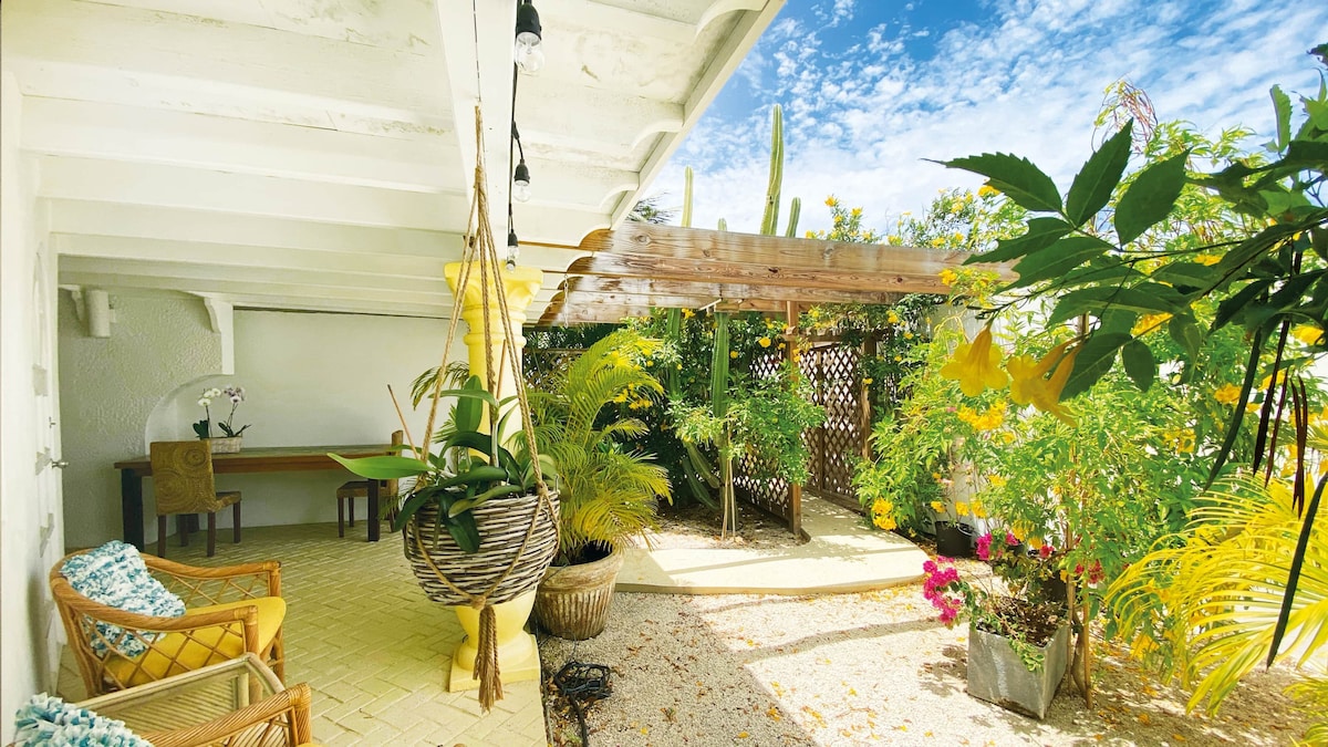 An outdoor patio area is shown, featuring a hanging chair and canopied seating. Lush greenery surrounds the space, complemented by colorful flower pots. Natural light filters through the trees, creating a serene atmosphere, while a wooden table and chairs are positioned in the background.