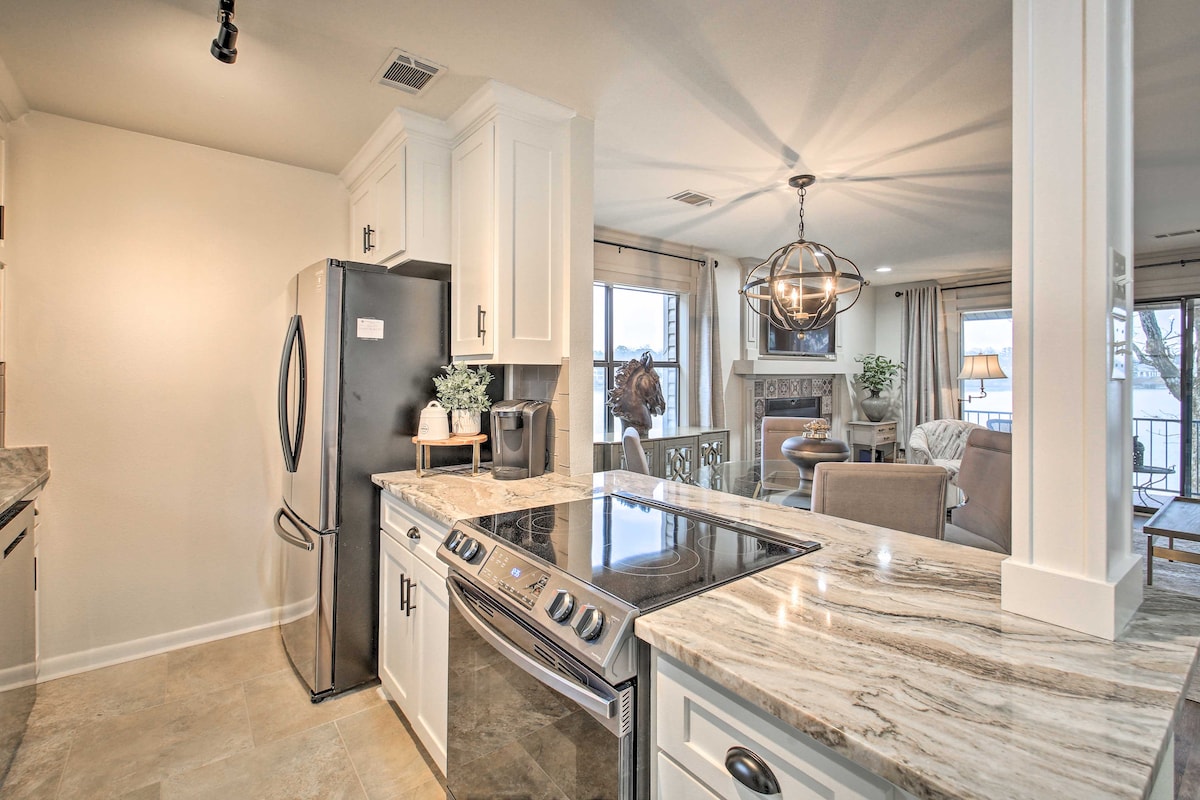 A modern kitchen features sleek white cabinetry and granite countertops. Stainless steel appliances include a refrigerator and stove, while a dining area with a pendant light is visible in the background. Natural light fills the space, highlighting the open layout towards the waterfront balcony.