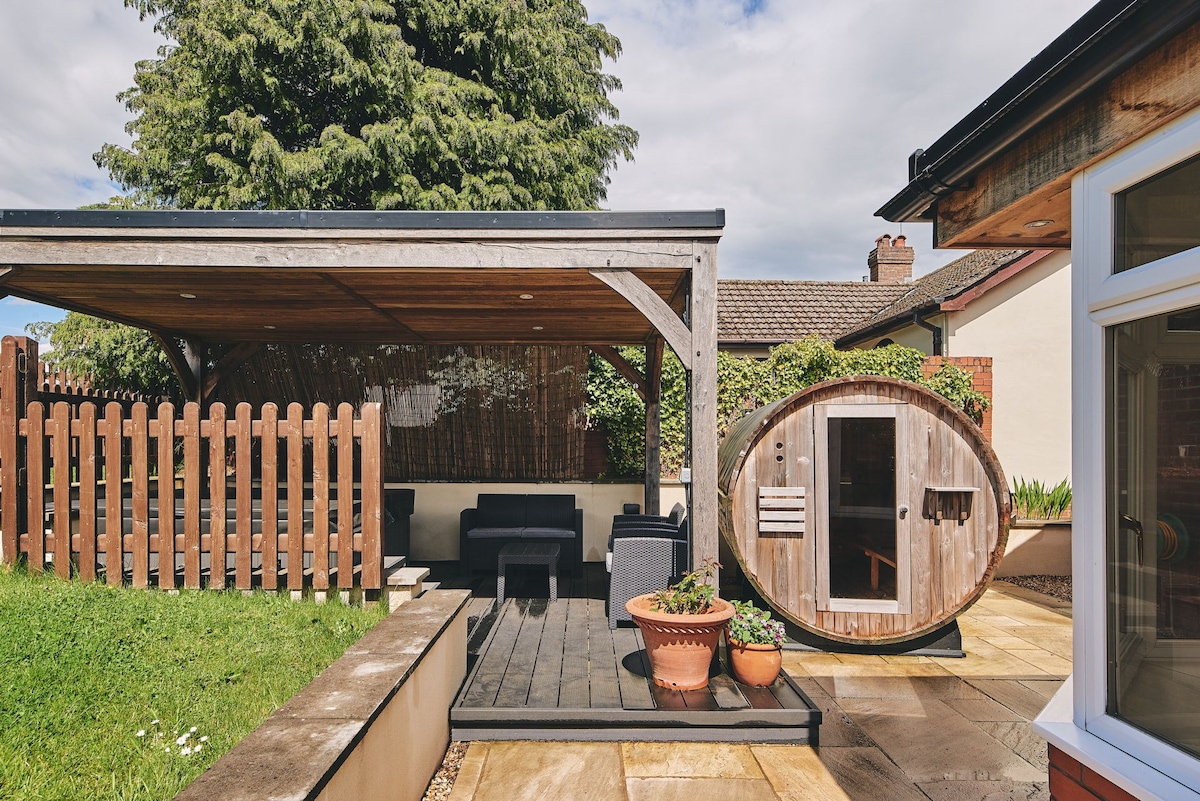 An outdoor area features a wooden structure providing shade and seating, accompanied by a barrel-shaped sauna. Potted plants and a well-maintained lawn enhance the space, while a wooden fence adds privacy, creating a serene environment for relaxation.