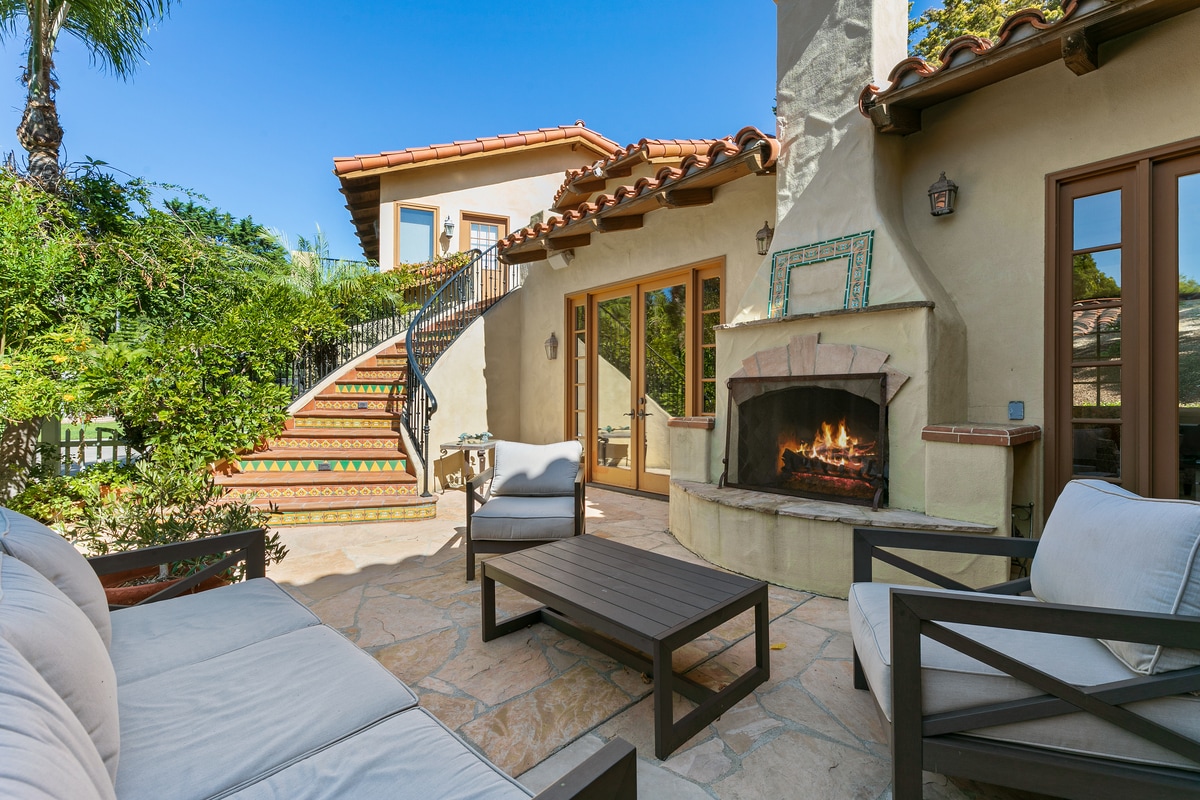 An inviting courtyard features comfortable seating around a stone fireplace, creating a cozy gathering space. Lush greenery surrounds the area, while stairs lead up to additional living spaces, highlighted by the warm glow of a fire. Sunlight bathes the scene in a relaxing ambiance.