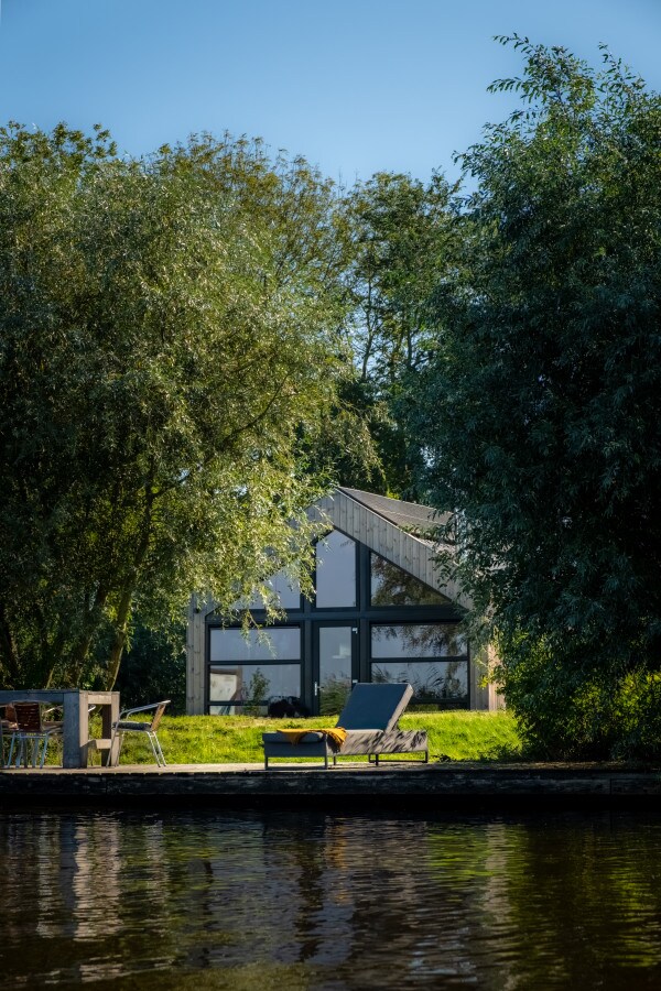 A modern nature house is nestled among tall trees, reflecting a serene environment. Sun loungers and a dining table are visible on a wooden deck by the water, inviting relaxation. Large glass windows showcase the bright interior, enhancing the connection with the surrounding greenery.