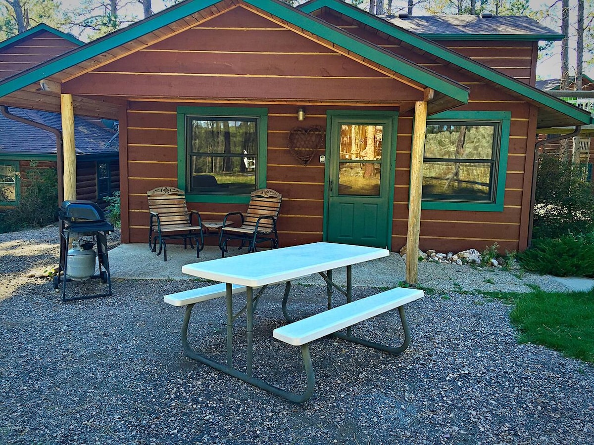 The cabin features a welcoming exterior with rustic wood siding in warm tones. A picnic table is positioned in front, accompanied by two chairs and a gas grill for outdoor dining. Lush greenery and gravel landscaping surround the cabin, enhancing the natural setting.