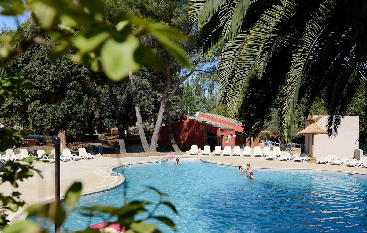 An outdoor swimming pool is surrounded by lush greenery and palm trees. Lounge chairs are arranged along the pool's edge, with a few guests enjoying the water. Nearby structures include a red building and another with a thatched roof.