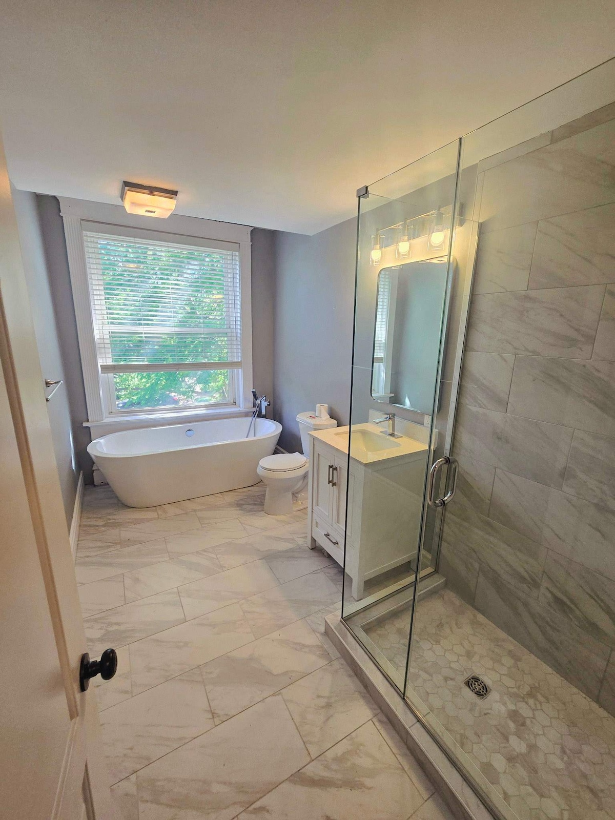 This modern bathroom features a spacious layout with a freestanding soaking tub and a walk-in shower. A white vanity provides storage, while natural light fills the space through a window, enhancing the elegant marble flooring and tiled walls.