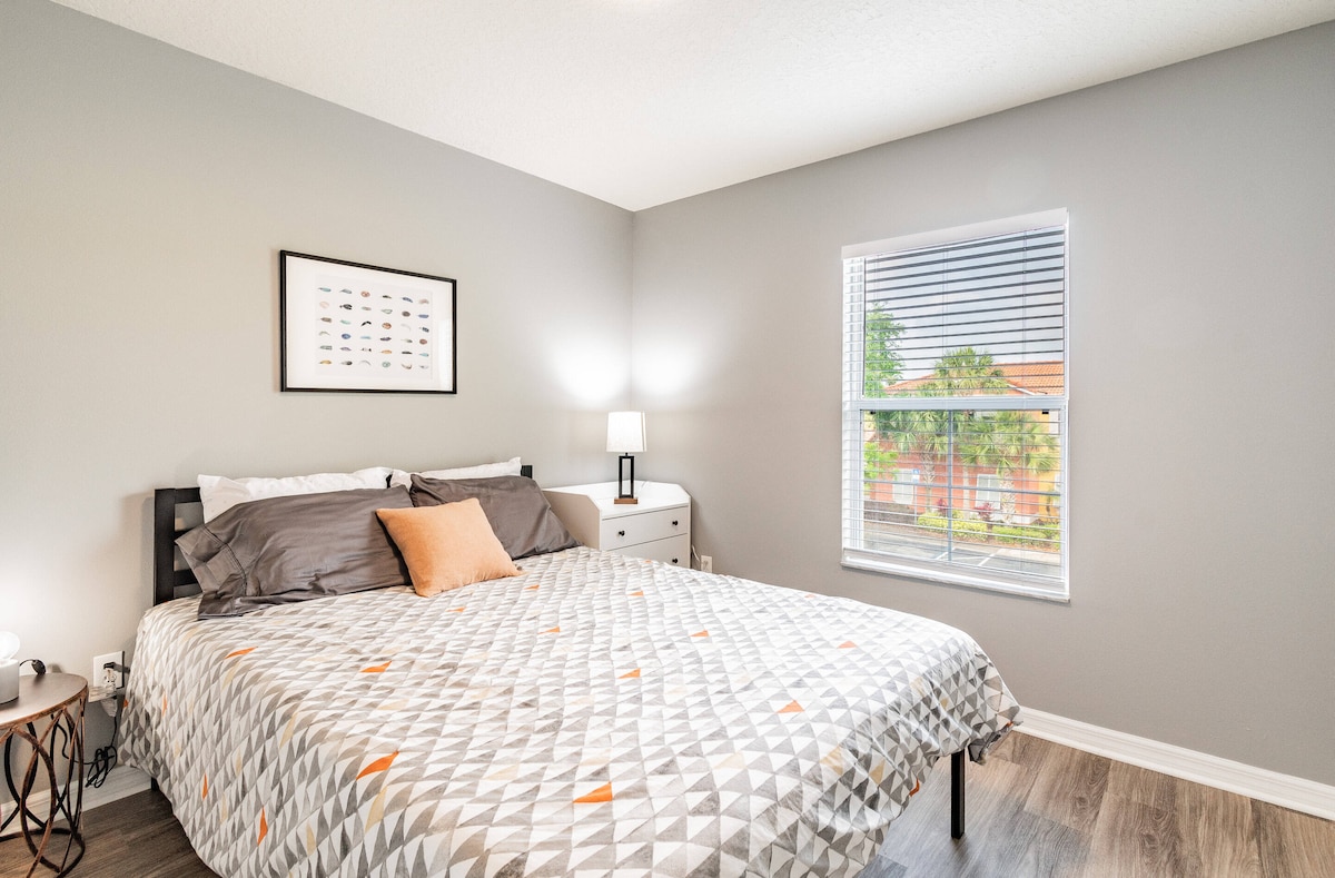 Ova svijetla i udobna spavaća soba ima elegantnu sivu paletu i topli drveni pod. Ugodna prošivena posteljina s jastukom u narančastoj boji unosi dašak boje, dok kroz prozor prodire prirodno svjetlo i pruža se lijep pogled na zelenilo.