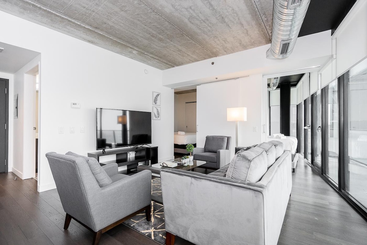 A modern living area features two grey sofas positioned around a glass coffee table, with a wall-mounted television offering entertainment options. Large floor-to-ceiling windows provide ample natural light, and the sleek flooring complements the industrial ceiling design.