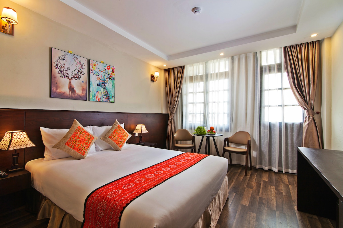 A well-appointed bedroom features a spacious bed with decorative pillows and a vibrant runner. Two artworks adorn the wall above the bed. Natural light filters through sheer curtains, complementing the warm tones of the wooden flooring and furniture.