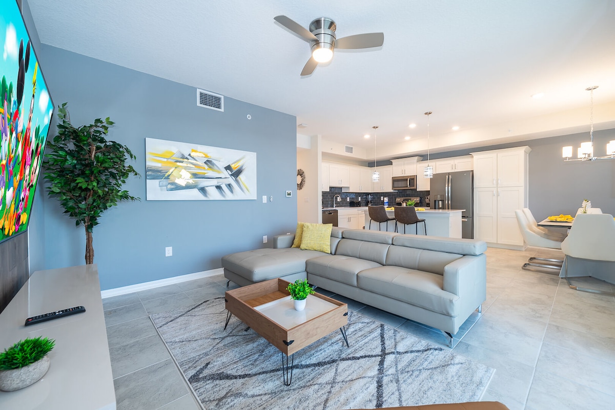 A modern living room showcases a light grey sectional sofa paired with a wooden coffee table. An expansive flat-screen television is mounted on the wall, and a vibrant piece of abstract art hangs nearby. The open-concept space allows for visibility of the dining area and kitchen.