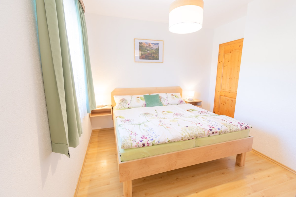 A spacious bedroom is presented, featuring a large bed with floral-patterned bedding. Soft lighting is provided by two bedside lamps. Natural light filters in through a large window with light green curtains, complementing the wooden bed frame and peaceful atmosphere.