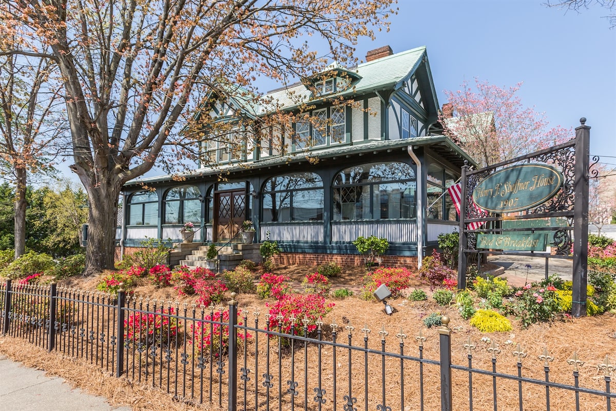 A charming two-story house is surrounded by vibrant floral landscaping and features a welcoming front porch with large windows. The exterior showcases a mix of green and wood accents, and an elaborately designed sign marks the entrance.