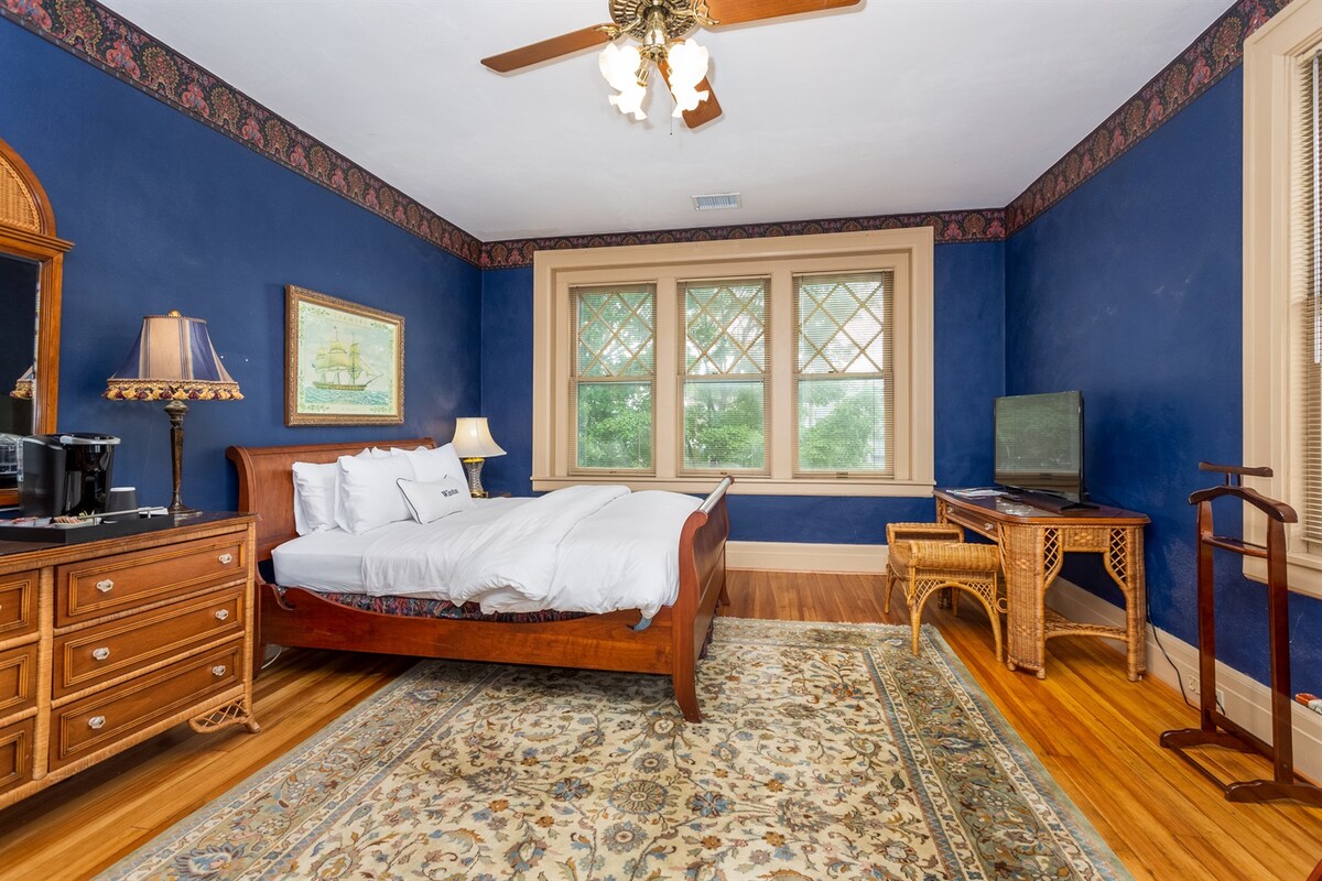 A spacious room features deep blue walls and a hardwood floor with a decorative area rug. A queen-size sleigh bed is positioned against one wall, accompanied by a wooden dresser and a wall-mounted flat-screen TV. Large windows provide natural light, and a ceiling fan is present.