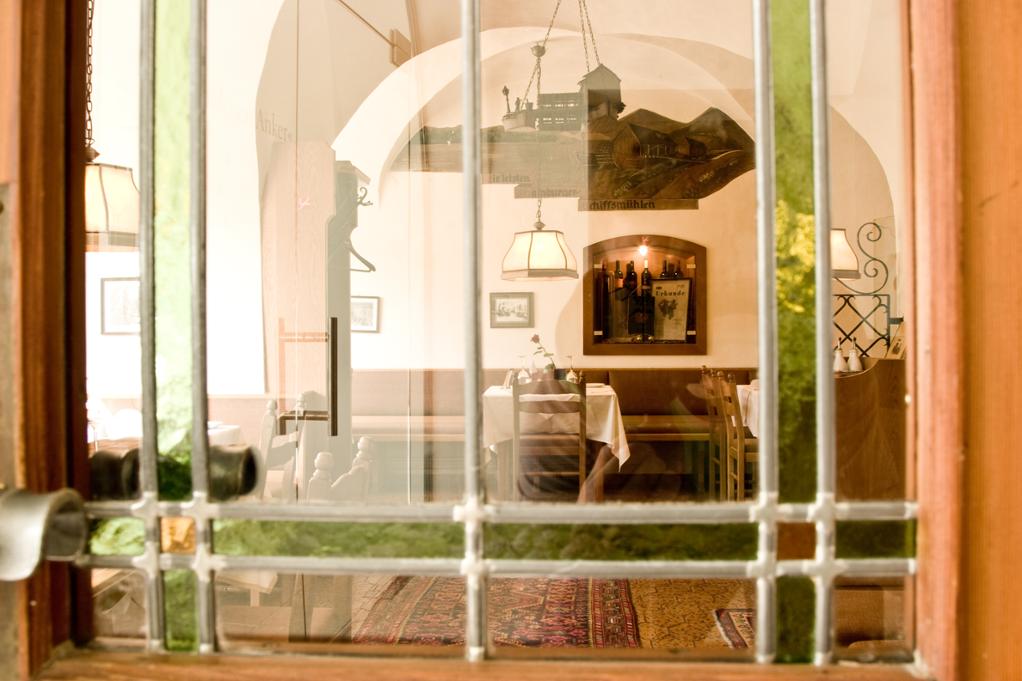 A view from outside shows a dining area within a cozy setting. Tables set with white linens are visible, framed by archways. Light fixtures cast a warm glow, while decorative elements adorn the walls, enhancing the inviting atmosphere of the space.