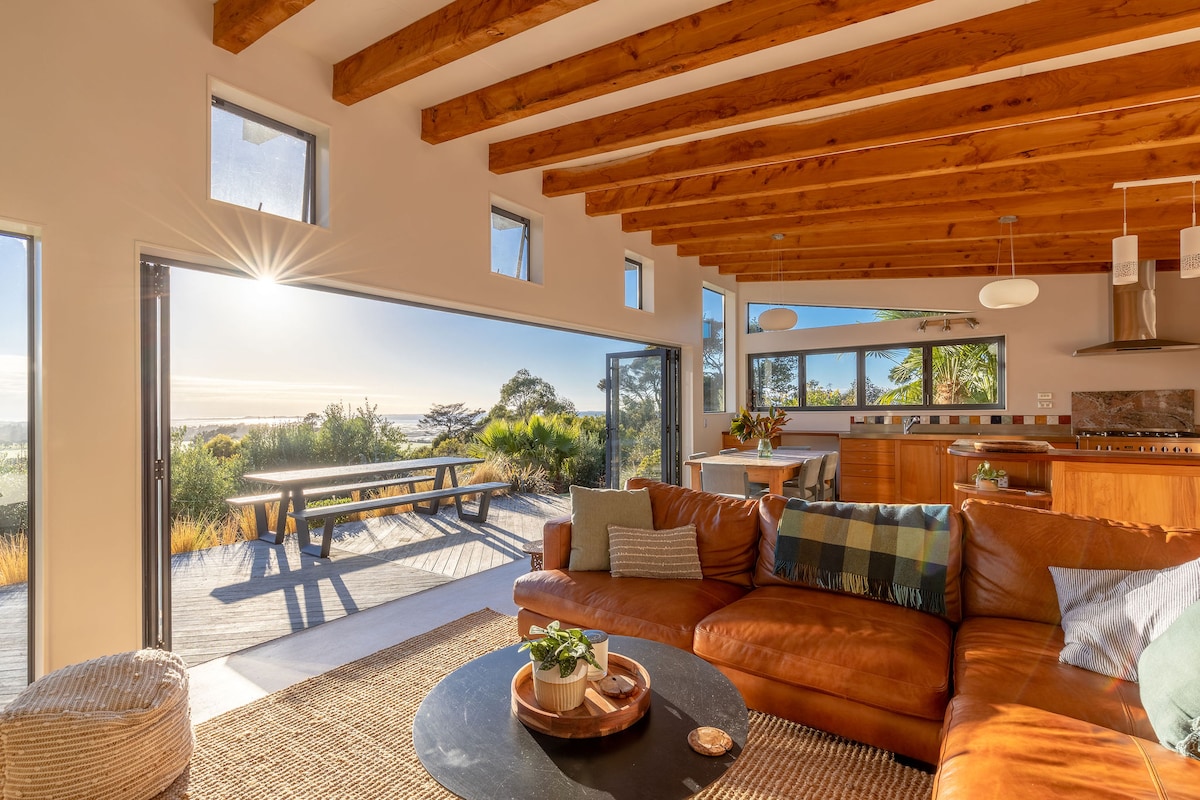 A spacious living area features a leather couch adorned with textured throw pillows. Large windows allow natural light to fill the space, highlighting wooden beams overhead. A round coffee table sits centrally, with an indoor plant adding a touch of greenery. The outdoor table is visible through glass doors.