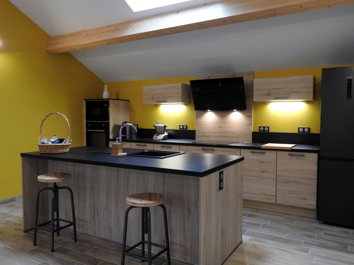 The kitchen features a central island with two wooden stools, surrounded by modern cabinetry in a warm wood finish. A black countertop provides a sleek surface, complemented by integrated appliances. A skylight allows natural light to brighten the space, while a vibrant yellow wall adds a touch of color.