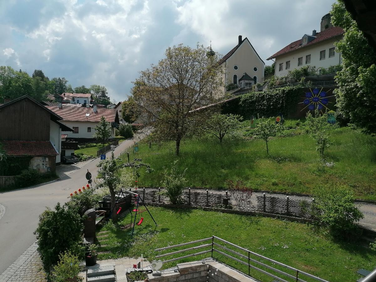 A scenic view captures a peaceful neighborhood, featuring green grass and a tree at the center. The road winds through the area, bordered by houses and small gardens. Buildings with varied architecture are seen on both sides, while a decorative feature stands out against the backdrop.