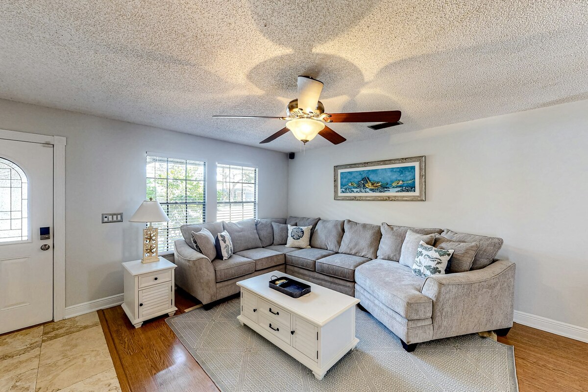 A cozy living room features a light-colored sectional sofa and a white coffee table. Natural light fills the space through multiple windows, and a ceiling fan adds comfort. A decorative lamp and wall art contribute to the welcoming ambiance.
