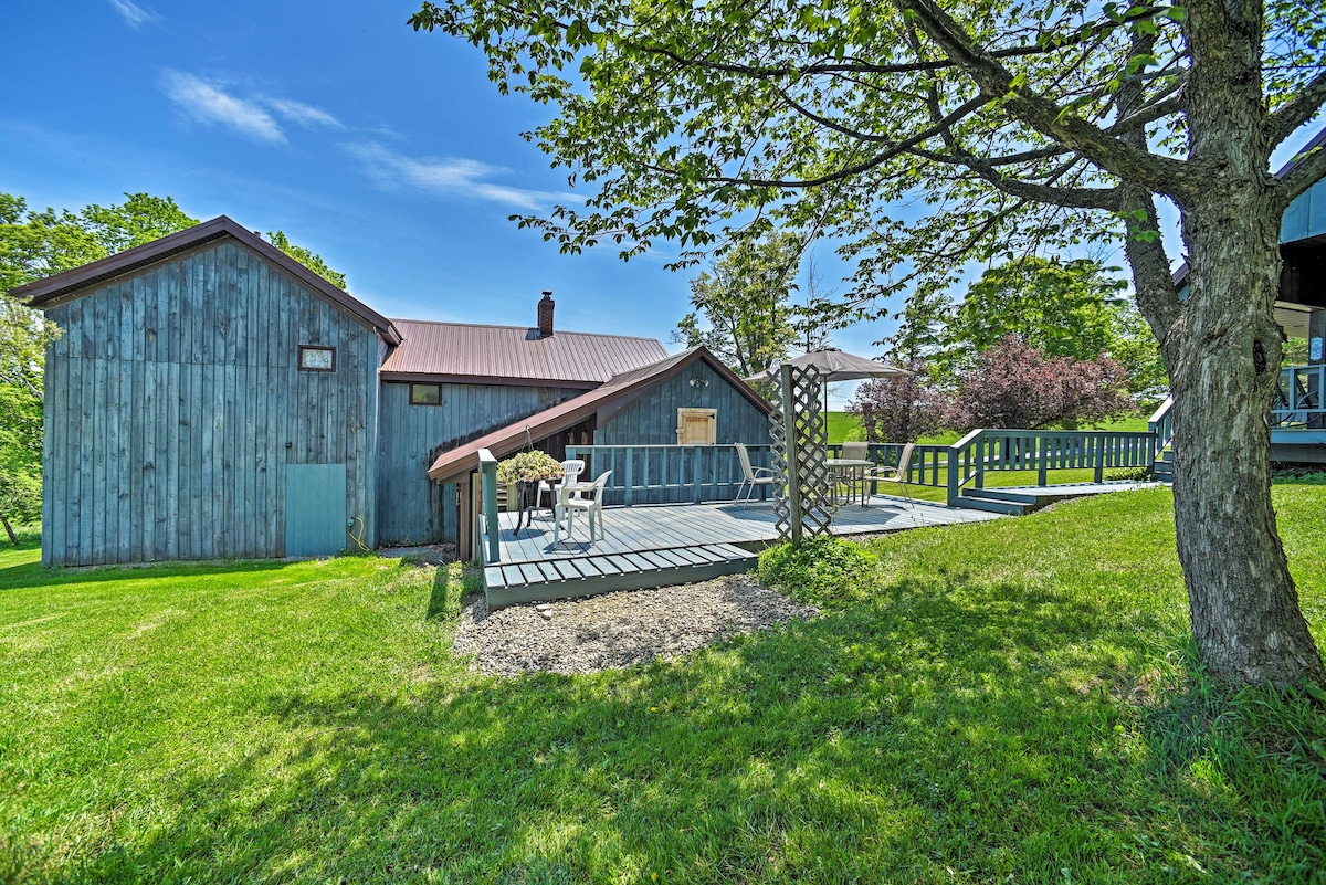 A rustic farmhouse exterior is viewed, featuring a wooden structure with a metal roof. A spacious deck leads to the entrance, surrounded by lush green grass and trees, offering a tranquil outdoor setting.