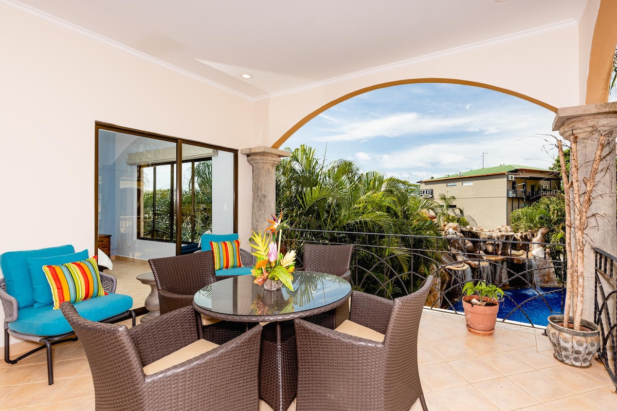 A terrace is furnished with a round dining table surrounded by six wicker chairs, providing a comfortable space for outdoor meals. A vibrant arrangement of cushions in blue and colorful stripes adorns two lounge chairs. Lush greenery and a pool are visible in the background.