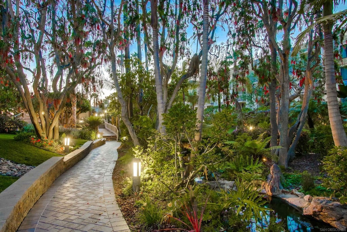 A winding stone pathway meanders through a lush garden, bordered by trees adorned with vibrant blossoms. Soft lighting accents the foliage, creating a serene ambiance. The pathway leads towards diverse greenery and a tranquil water feature, inviting exploration in a peaceful outdoor setting.