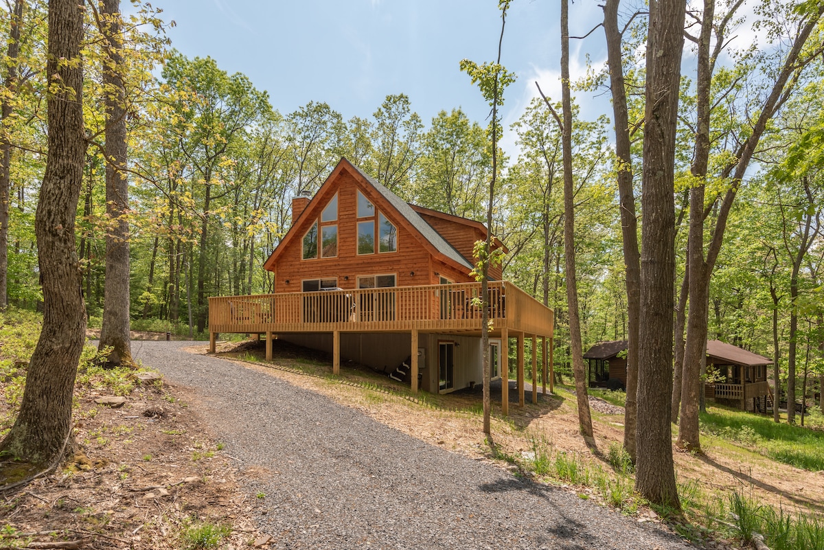 The cabin features a large wrap-around deck, with wooden railings and steps leading to the entrance. Surrounded by trees, the two-story structure showcases a blend of wood and modern design elements. A gravel driveway provides access to the property, enhancing the natural setting.