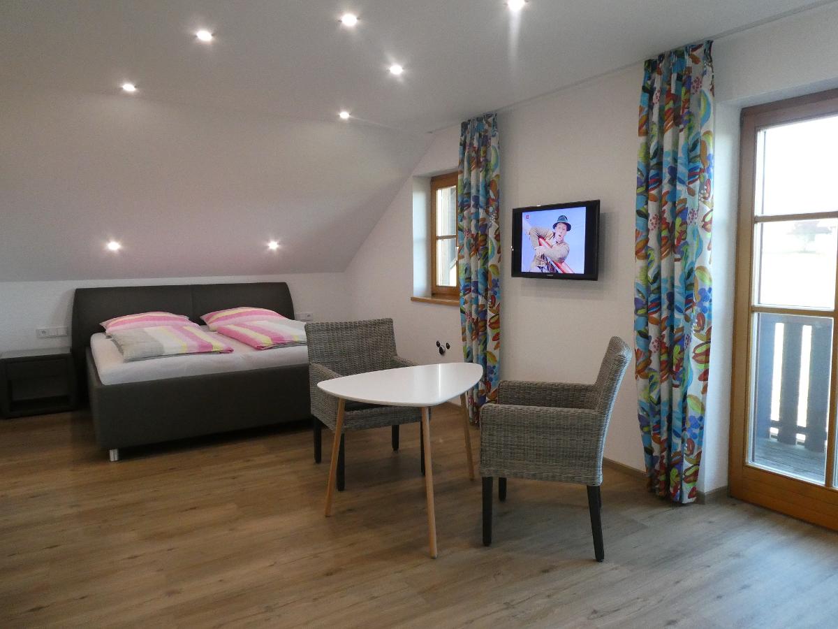 A comfortable room features a spacious bed with colorful bedding. A small round table sits between two woven chairs, while a flatscreen TV is mounted on the wall. Large windows are framed by patterned curtains, allowing natural light to fill the space.