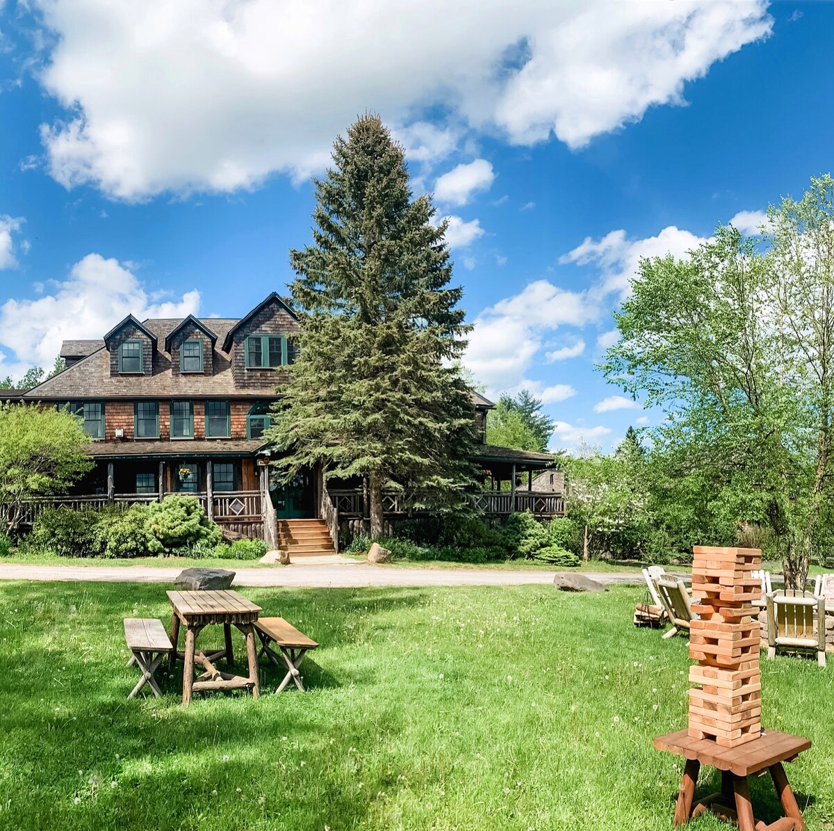 A large lodge is set against a backdrop of blue skies and fluffy clouds. Lush green grass surrounds the building, which features multiple stories and a balcony. Wooden picnic tables are arranged on the lawn, alongside a stacked game of outdoor Jenga.