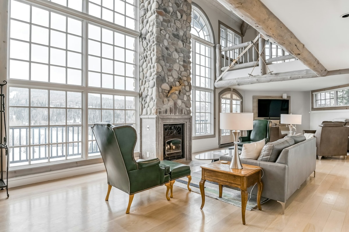 A spacious living area features large windows that provide ample natural light and a view of the mountains. A stone fireplace is accented by decorative elements, while comfortable seating options include a green wingback chair and a grey sofa. A wooden coffee table sits in the center.