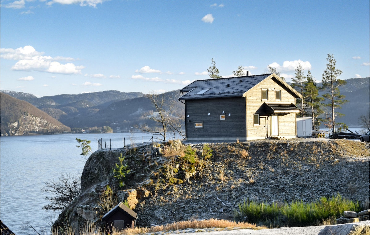 A two-story holiday home is situated on a cliff overlooking the water, surrounded by natural scenery. The building features a combination of wooden and modern materials, with a small outdoor deck visible. Nearby trees and rolling hills create a tranquil backdrop.