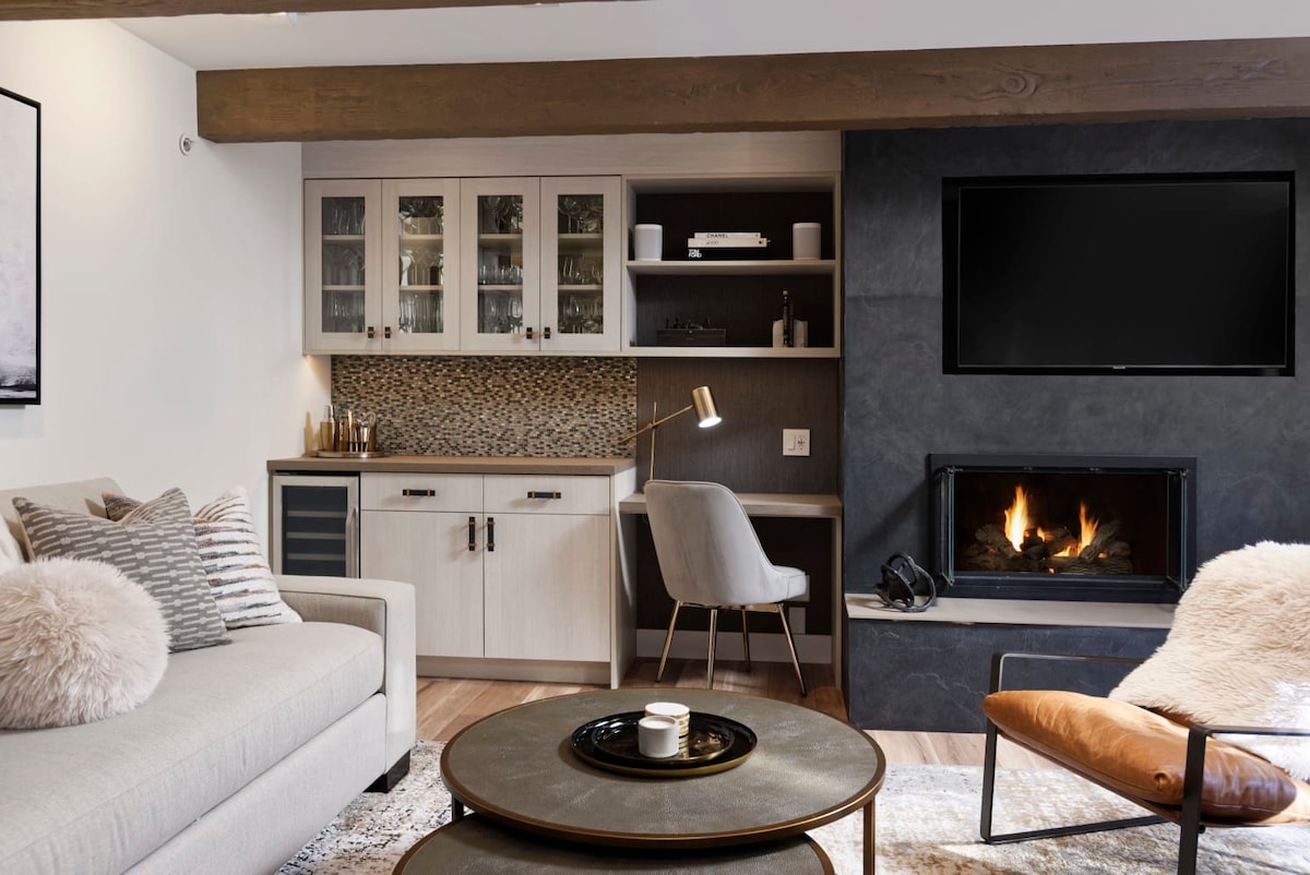 A stylish living area is showcased with a light-colored sofa, accented by plush pillows. A modern coffee table sits in front, while a wood-burning fireplace displays a warm glow. Built-in cabinets and a dedicated workspace enhance functionality, with a wall-mounted TV positioned nearby.