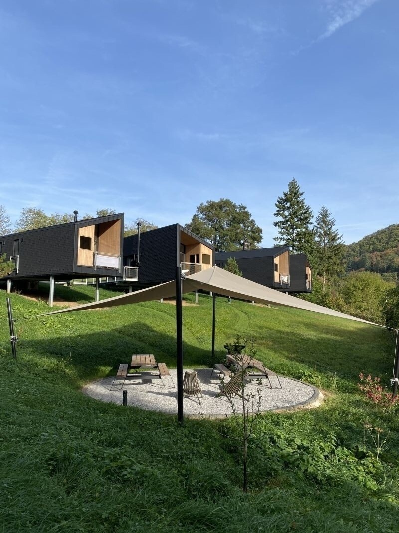 Three minimalist lodges are positioned on a gently sloping terrain, nestled among greenery. A shaded seating area with a picnic table and a circular stone pattern enhances the natural surroundings, while a large tarp provides shelter. The blue sky and trees form a serene backdrop.
